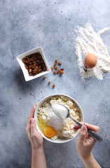 Cooking process. Preparation. Cottage cheese in a white bowl, raisins, eggs and flour on a light grey background with hands. Flatlay, top view. Background image, copy space