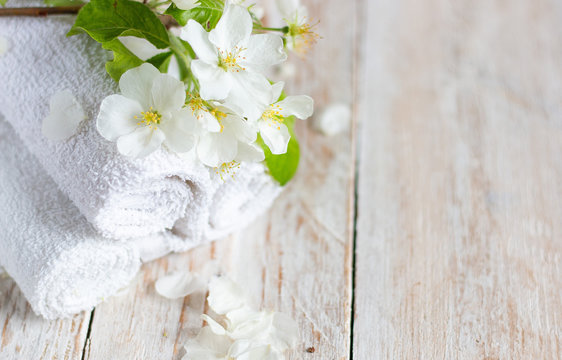 Three white towels rolled up, flowering branch with white flowers on a light wooden table. Spa and beauty treatment concept. Copy space for text.