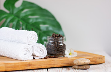 Handmade coffee scrub in a glass jar on a light wooden table. White towels rolled up. Homemade peeling and spa care product.