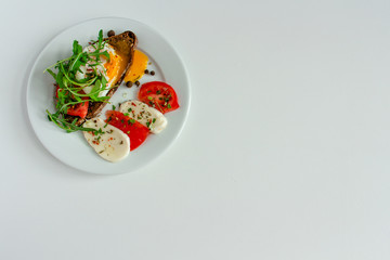delicious poached egg on a slice of black rye bread with slices of tomato, mozzarella, arugula, spices and black pepper on a white plate on a white background