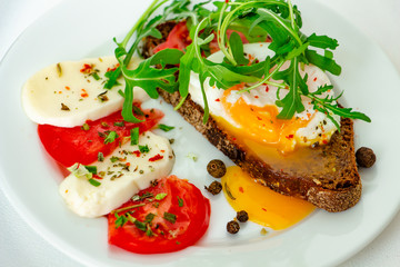 delicious poached egg on a slice of black rye bread with slices of tomato, mozzarella, arugula, spices and black pepper on a white plate on a white background