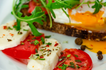 delicious poached egg on a slice of black rye bread with slices of tomato, mozzarella, arugula, spices and black pepper on a white plate on a white background