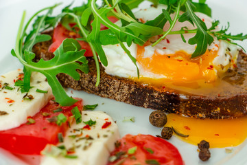 delicious poached egg on a slice of black rye bread with slices of tomato, mozzarella, arugula, spices and black pepper on a white plate on a white background