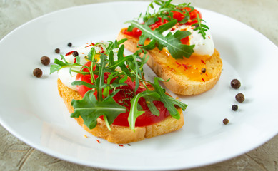 delicious poached eggs on crispy slices of wheat bread with slices of tomato, arugula, spices and black pepper on a white plate on a concrete background