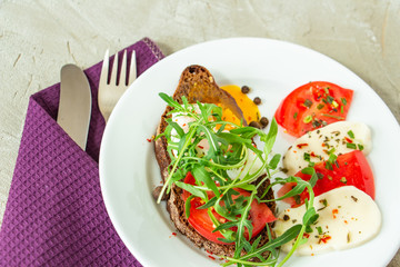 delicious poached egg on a slice of black rye bread with slices of tomato, mozzarella, arugula, spices and black pepper on a white plate on a concrete background