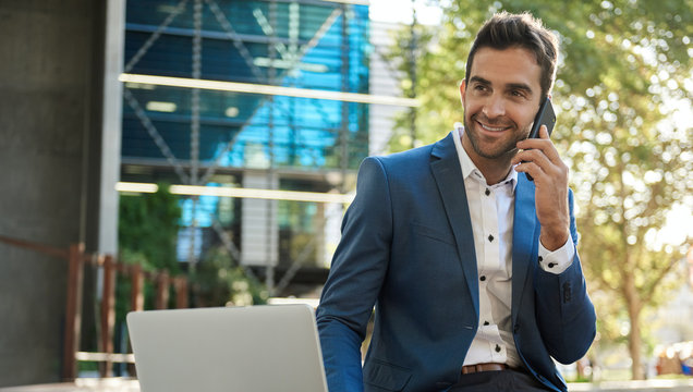 Smiling Businessman Sitting Outside Using A Cellphone And Laptop