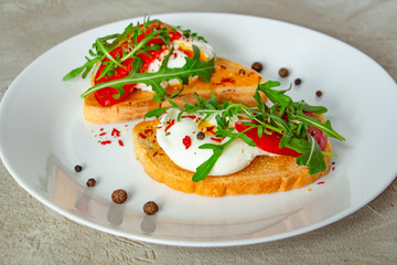 Fototapeta na wymiar delicious poached eggs on crispy slices of wheat bread with slices of tomato, arugula, spices and black pepper on a white plate on a concrete background
