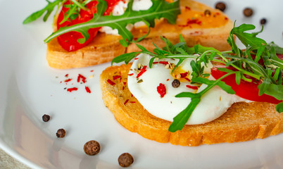 delicious poached eggs on crispy slices of wheat bread with slices of tomato, arugula, spices and black pepper on a white plate on a concrete background