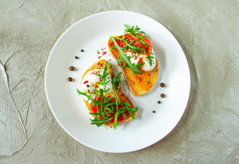 delicious poached eggs on crispy slices of wheat bread with slices of tomato, arugula, spices and black pepper on a white plate on a concrete background