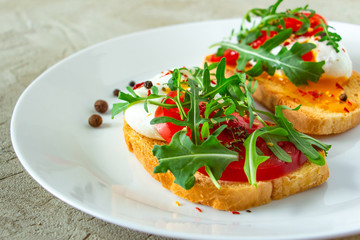 delicious poached eggs on crispy slices of wheat bread with slices of tomato, arugula, spices and black pepper on a white plate on a concrete background