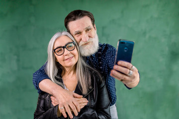 Fototapeta premium Smiling satisfied senior couple looking together at phone camera for taking selfie photo, posing on green wall background, smiling and embracing