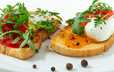 Fototapeta na wymiar delicious poached eggs on crispy slices of wheat bread with slices of tomato, arugula, spices and black pepper on a white plate on a white background 