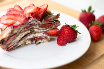 Light summer dessert: a slice of crepe layer cake with whipped cream, chocolate chips and strawberries between the layers. It is decorated with strawberries and placed on a white plate.