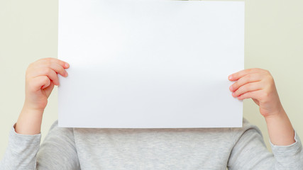 Blank paper in hands of child covering her face on light background. Mockup white paper in child's hands. Empty space for text.