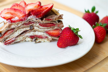 Light summer dessert: a slice of crepe layer cake with whipped cream, chocolate chips and strawberries between the layers. It is decorated with strawberries and placed on a white plate.