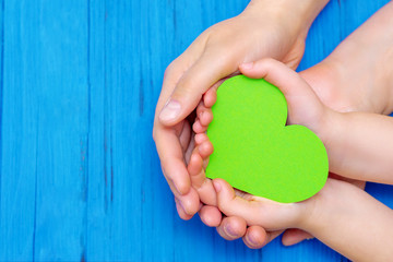 Obraz premium Top view of child and father hands holding paper green heart on wooden blue background. Concept of family and family relations.