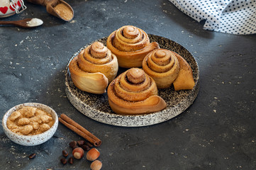 Cinnabon rolls on a ceramic plate next to a bowl with brown sugar, cinnamon sticks and nuts. Homemade cakes in the shape of a snail with ingredients. Textured background and plate..