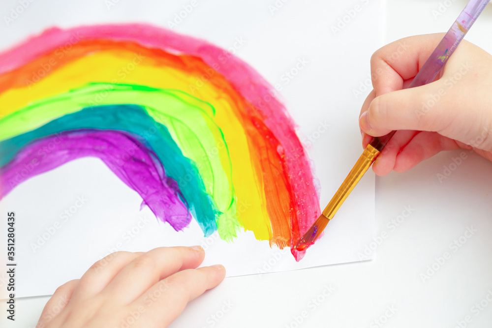 Wall mural Close up of hands of child drawing rainbow by watercolors on white paper.