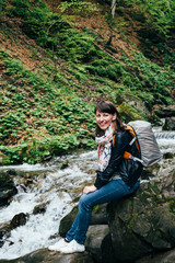 Naklejka na ściany i meble Happy smiling tourist girl in blue jeans, striped shirt and black jacket with backpack near to the cold fresh river at the waterfall Shypit, cascade in Pylypets in the forest. Carpathian Mountains