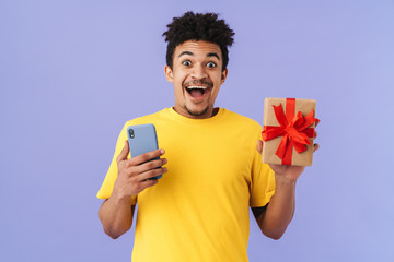 Photo of astonished african american man holding gift box and cellphone