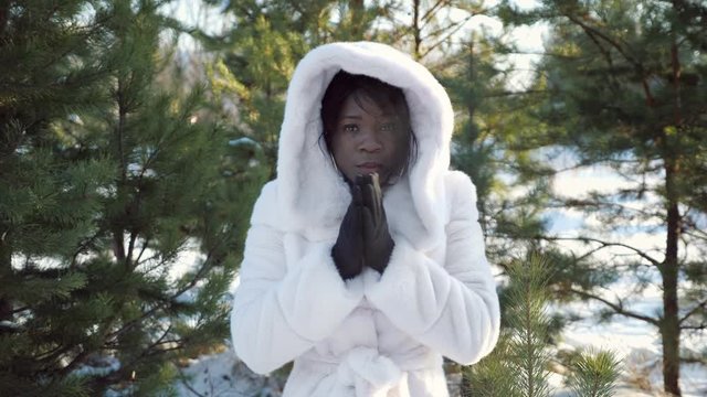 sad dark-skinned girl in hood trembles with cold and warms hands against green pine-trees in cold winter closeup