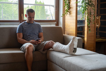 Man with broken leg in cast using mobile phone while sitting in sofa at home