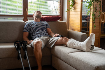 Side view of a young handsome bearded man in casual clothes sleeps on the couch in a room at home. with feet in plaster