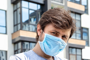 Fototapeta na wymiar Portrait of a bearded man in protective blue medical mask against a blurred outdoor house with balconies background. Male in a respirator to protect against infection with coronavirus or smog