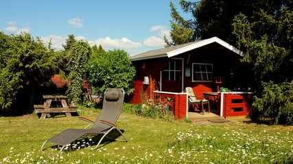 natürlicher erholsamer Garten mit Margeriten und rotem Gartenhaus und Liege unter Bäumen