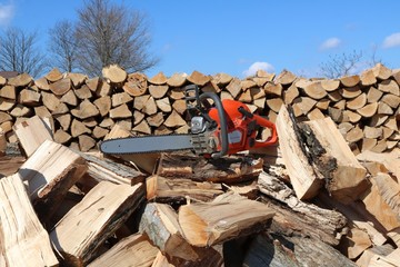 Orange and grey chainsaw on top of heap of wood with split piled wood behind and blue sky