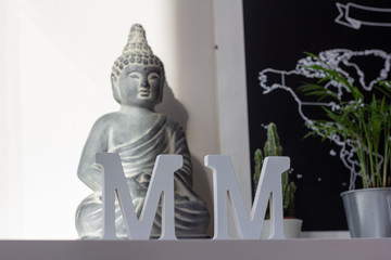 Two white decorative letters M with blurred background with a plant, a world map and a stone Buddha