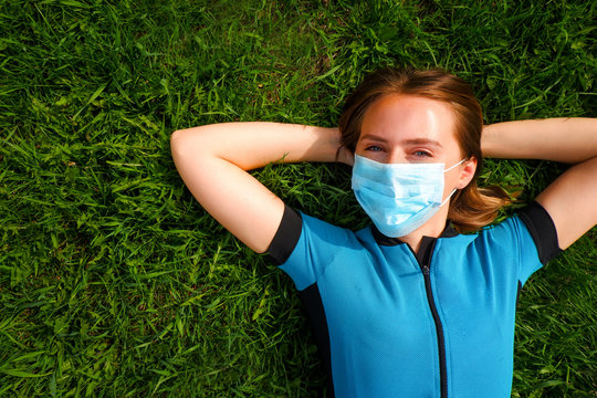 A young woman  of European appearance in blue in a protective medical mask lies on green grass in the park during quarantine covid-19. Spring Summer.  The girl look into the frame.
