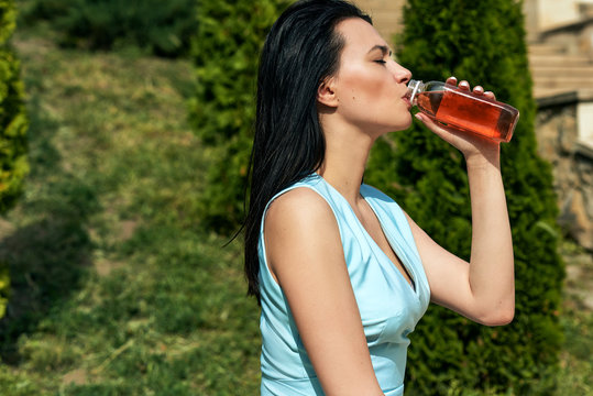 Outdoor side view image of fitness young woman drinking fresh juice from the bottle. Stylish girl drinking detox drink in a glass jar in the street. Caucasian female model taking a cleanse diet.