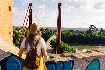 Girl with cap, sunglasses, golden sports jacket and leather backpack facing the river at dawn ....