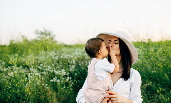 A horizontal outdoor image of pretty mother and cute little girl hug each other, on the field, on sunset sunlight. Mom and daughter sitting on a field of flowers on nature background.