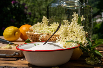 On the wooden table there is an enamel bowl with sugar, a vanilla pod and a basket with fish harvested elderberry blossoms to make a liqueur