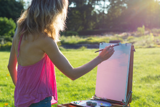 A young woman artist holds a brush and paints a picture on an easel iof the sunset.
