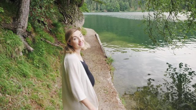 Back view of happy pregnant woman walking near mountain lake.