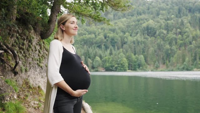Pregnant woman touching gently her tummy when standing near the mountain lake. Walking of pregnant woman at nature