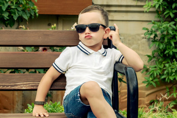 A cool child in sunglasses sits on a bench with his legs crossed. A handsome boy of 4 years old...