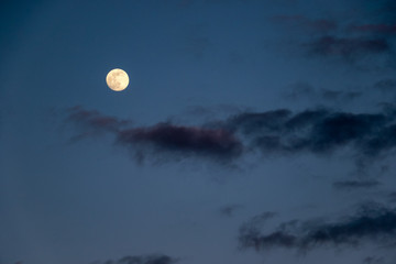 A bright round full moon in the night sky surrounded by wispy clouds.