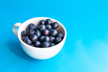 Ripe blueberry and green leaves in white cup on blue horizontal background