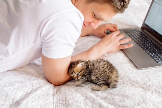 Young Handsome Man Working At Home On Self-isolation While Quarantining A Coronovirus, Lying With A Laptop And A Kitten In Bed