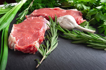 Raw meat and fresh herbs. Two slices of fresh raw meat, a branch of rosemary and garlic on a black...