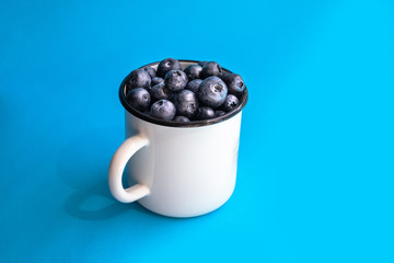 Minimal composition of fresh blueberries in a white metal mug on a blue background. Healthy eating