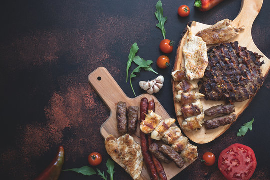Various grilled meats served on wooden cutting boards with fresh vegetables and bread on a dark rustic background. Traditional Serbian and Balkan bbq meat called rostilj. Flat lay. Copy space