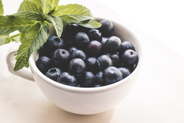 Ripe blueberry and green leaves in white cup on a light background
