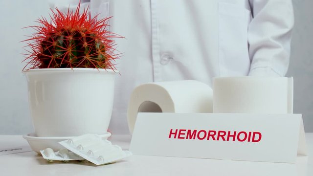 Doctor putting paper with word HEMORRHOID on table with cactus, suppositories and toilet paper