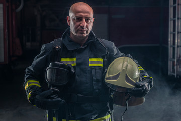 Fototapeta premium Fireman standing confident holding helmet and wearing firefighter turnouts. Portrait of a fireman with dark background with smoke and blue light.