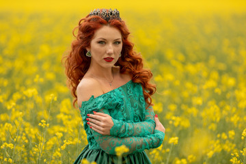 Beautiful Redhead female model in a field of yellow rapeseed flowers. Wearinf at long green dress and crown